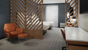 A hotel room with a wooden partition, a single bed, a desk, an orange armchair, and a small round ottoman.