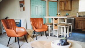 Un salon chaleureux avec deux fauteuils en cuir, une petite table ronde et un canapé. On aperçoit un coin cuisine avec des meubles en bois et, au fond, une porte de couleur turquoise.