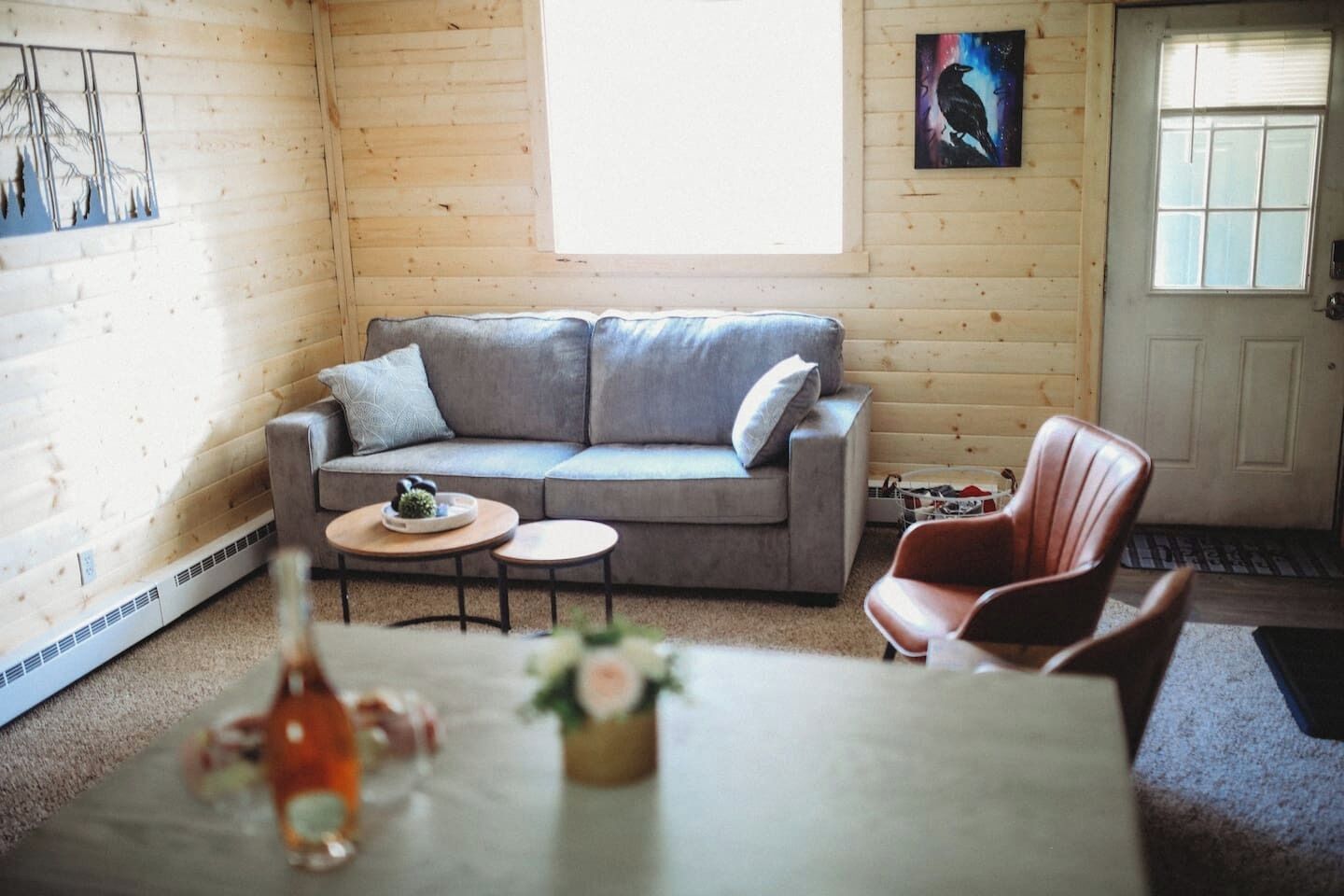 A cozy room with a grey sofa, two small round tables, a wooden floor, a door, and a window.