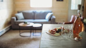 Une table à manger avec une bouteille, des verres et une assiette de nourriture, dans une pièce où se trouvent un canapé et une petite table ronde.