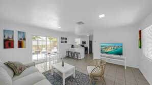 A modern living room with a white sofa, a coffee table, and a TV on a stand. There is a kitchen area in the background with a dining table and chairs. - Heart of Calle 8 | Spacious 2/2 for Families (Miami)