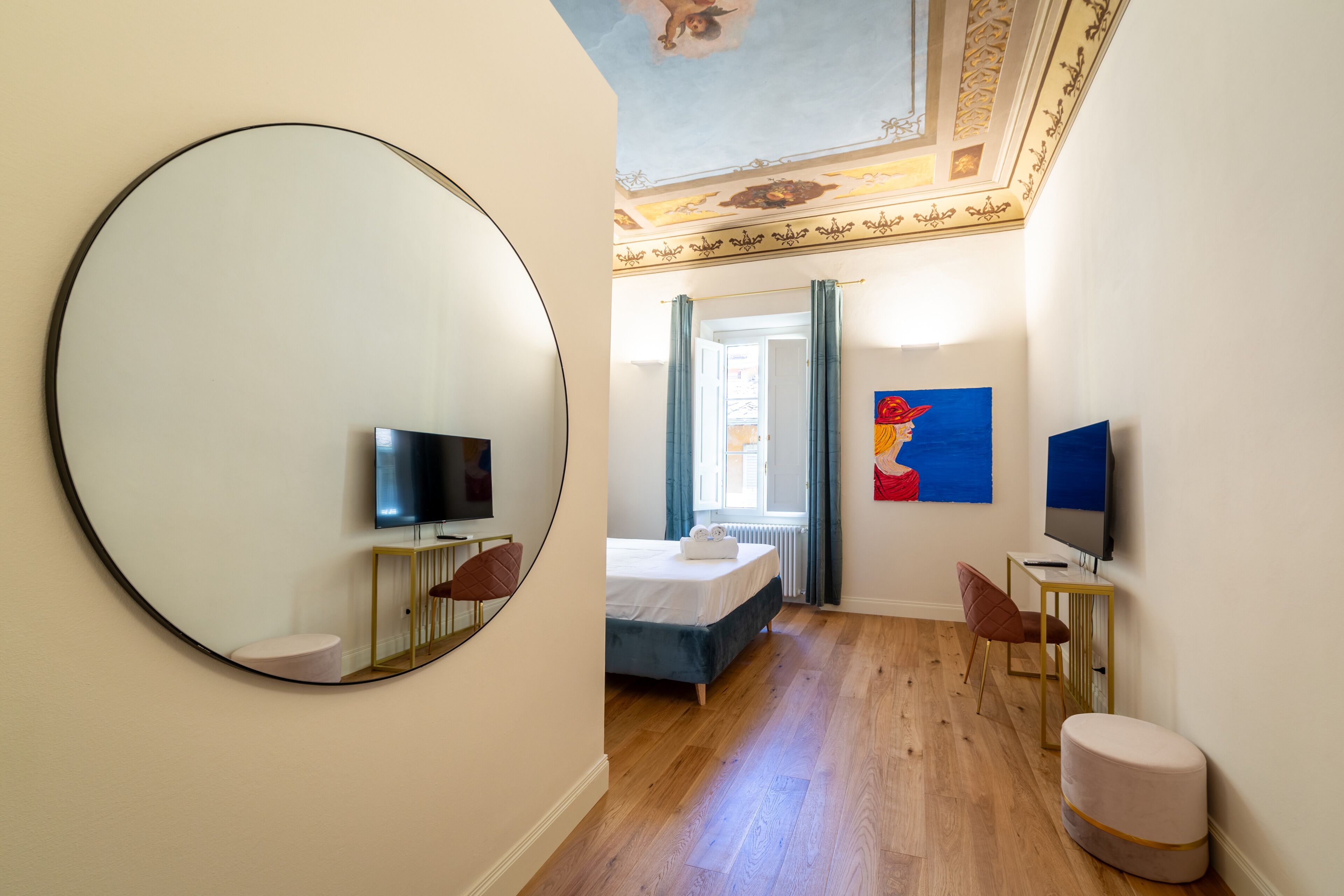 Una camera d'hotel con un ampio specchio rotondo, un letto, una scrivania con una sedia, una televisione e un quadro appeso al muro.