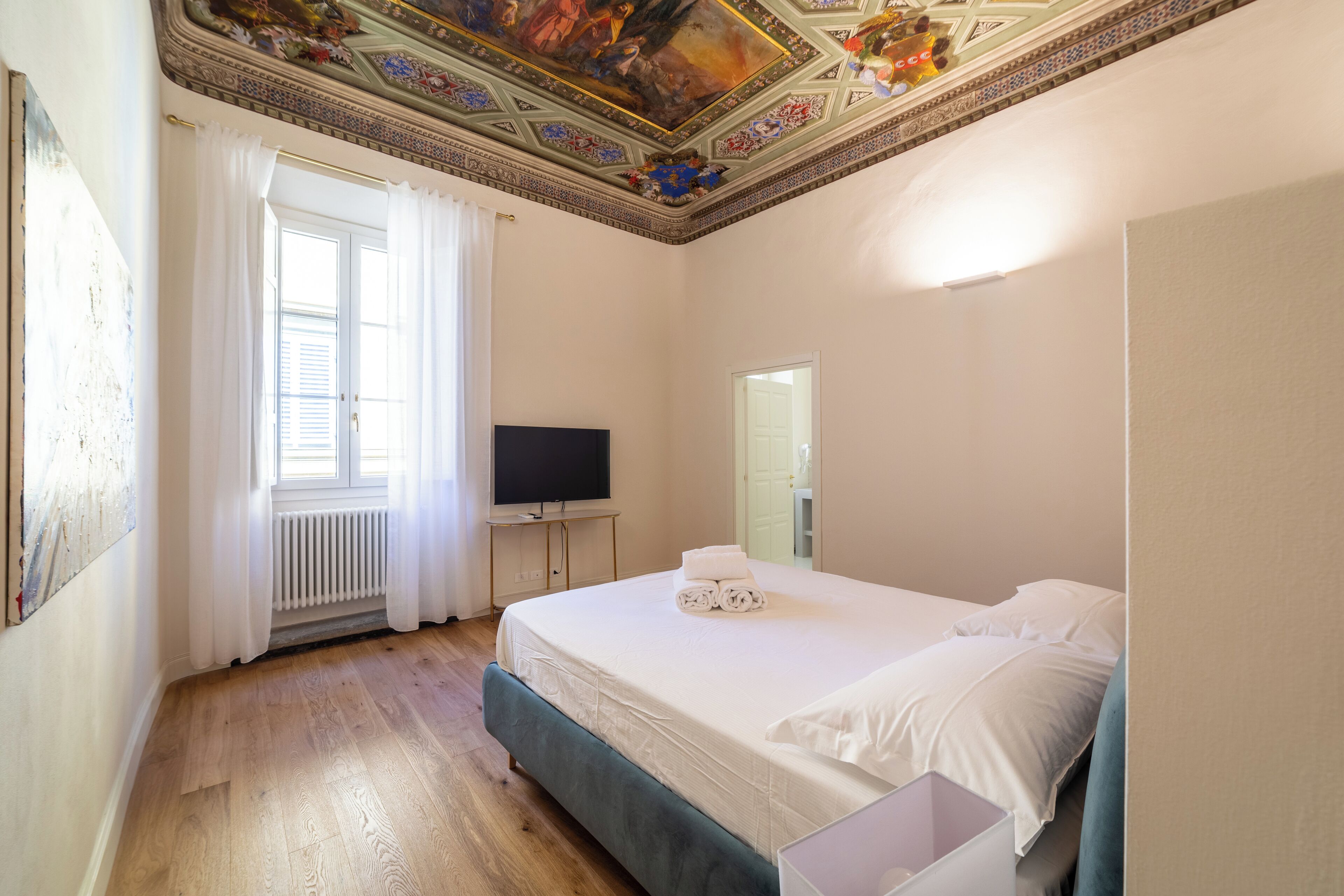 Un dormitorio con una cama grande, un televisor sobre un mueble, una ventana con cortinas y un techo decorativo.