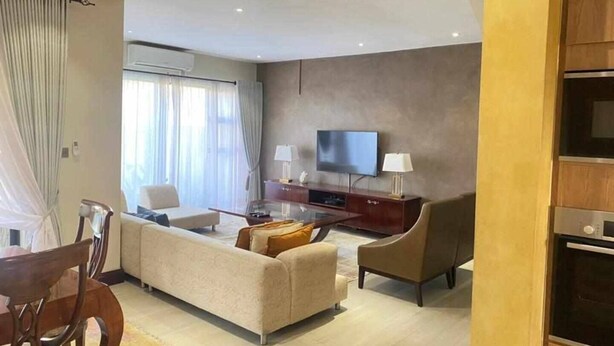 A modern living room with a sofa, chairs, a TV, and a dining area. - Roma Park 2 Bedroom Villa (Lusaka)