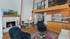 A spacious living area with a fireplace, a dining table with chairs, a kitchen with wooden cabinets, and a balcony with a view. - Summit View Treehouse By Destination Big Bear: Pet Friendly! Close to Bear Mtn! Walking Distance to Alpine Zoo! (Big Bear Lake)