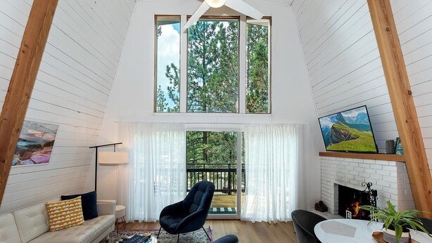 A cozy living room with a high ceiling, a fireplace, a dining area, and large windows offering a view of trees. - Summit View Treehouse By Destination Big Bear: Pet Friendly! Close to Bear Mtn! Walking Distance to Alpine Zoo! (Big Bear Lake)