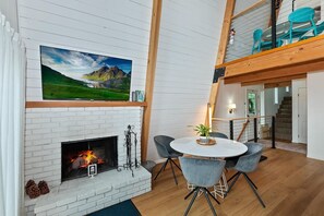 A modern living room with a fireplace, a television mounted above it, and a dining area with chairs. - Summit View Treehouse By Destination Big Bear: Pet Friendly! Close to Bear Mtn! Walking Distance to Alpine Zoo! (Big Bear Lake)