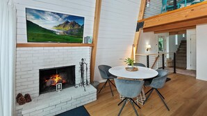 A modern interior with a fireplace, a dining area with a round table and chairs, and a large TV mounted on the wall. - Summit View Treehouse By Destination Big Bear: Pet Friendly! Close to Bear Mtn! Walking Distance to Alpine Zoo! (Big Bear Lake)
