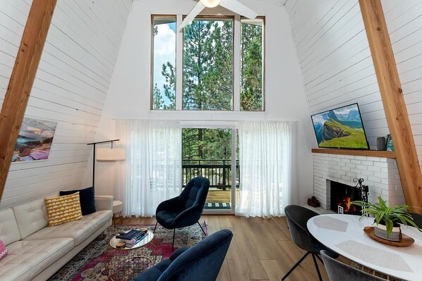 A high-ceilinged room with a fireplace, a sofa, a dining table, and a view of trees through large windows.