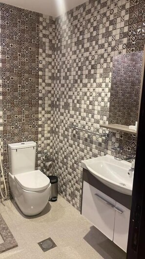 A bathroom with a patterned tile wall, a toilet, a trash can, and a sink with a mirror.