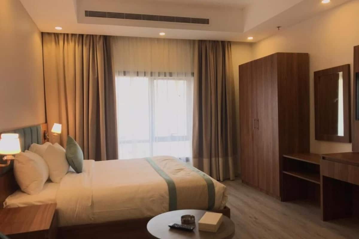A hotel room with a large bed, a desk, a chair, a small table, a wardrobe, and a window with curtains.