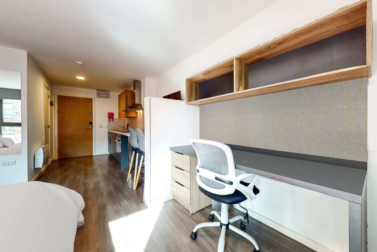 A modern hotel room with a wooden floor, a desk with a chair, a bed, and a kitchenette in the background.