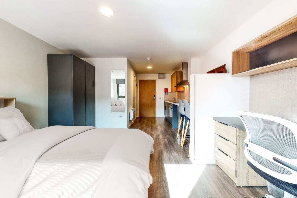 A modern hotel room with a large bed, a desk with a chair, a kitchenette, and a bathroom visible through an open door.