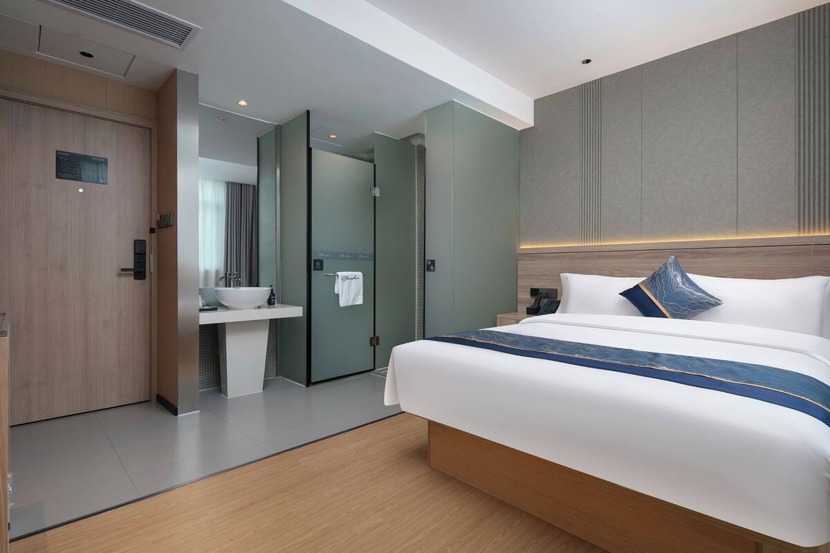 A modern hotel room with a large bed, a bedside table, a built-in TV, and a bathroom with a sink and mirror.