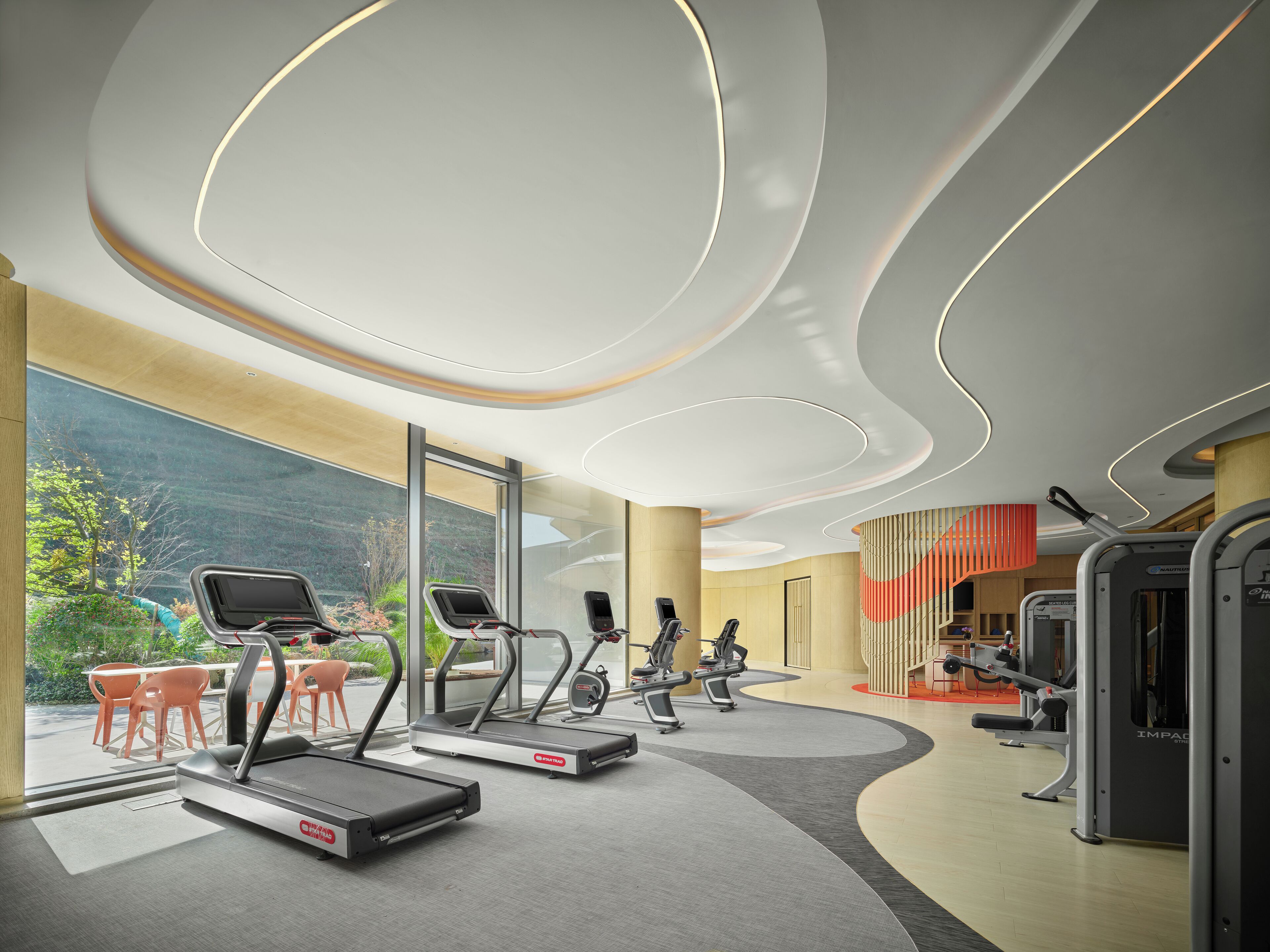 A modern gym with treadmills, exercise equipment, and large windows offering a view of greenery.