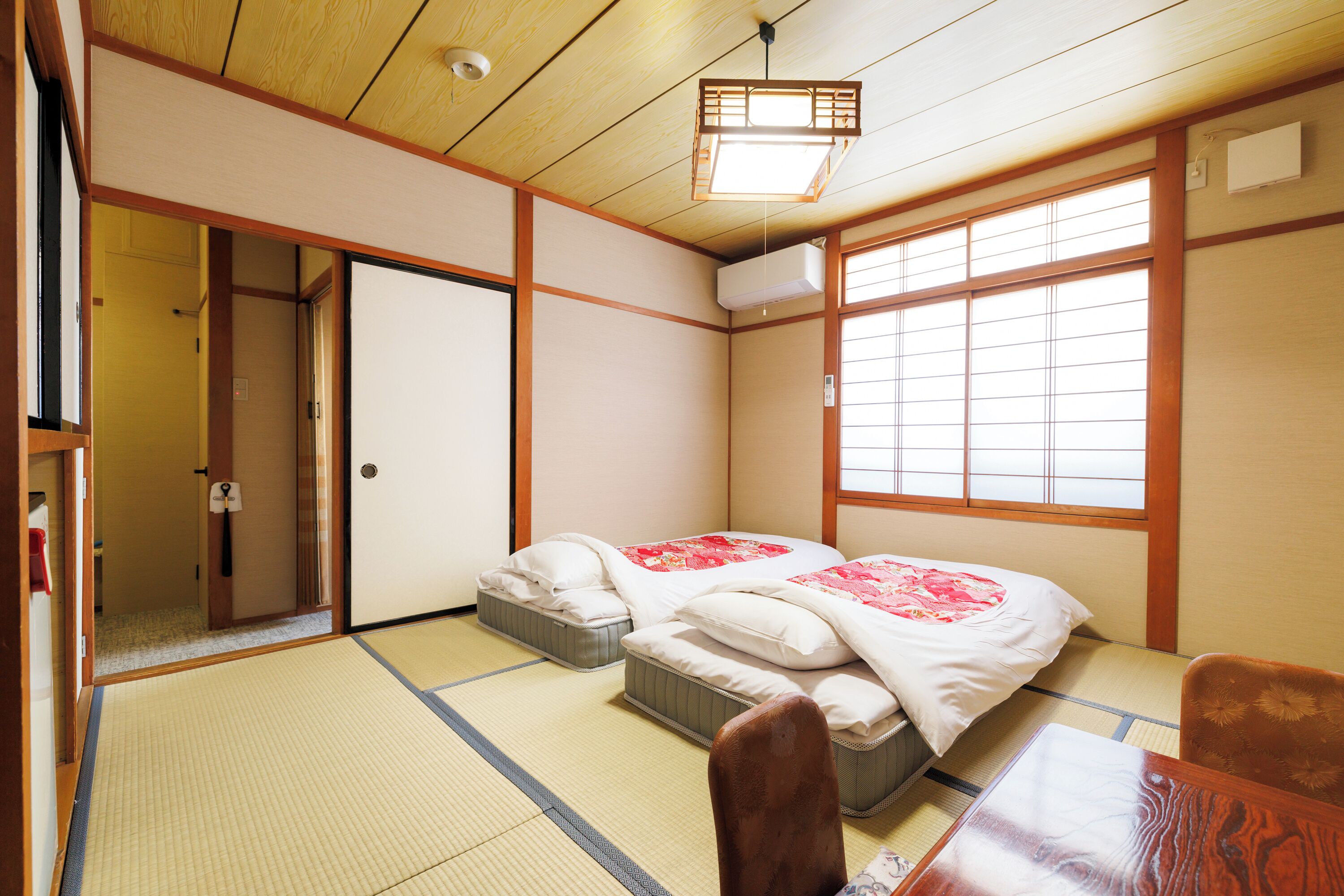 A traditional Japanese room with tatami flooring, a low wooden bed with red bedding, a wooden table, and a sliding door.