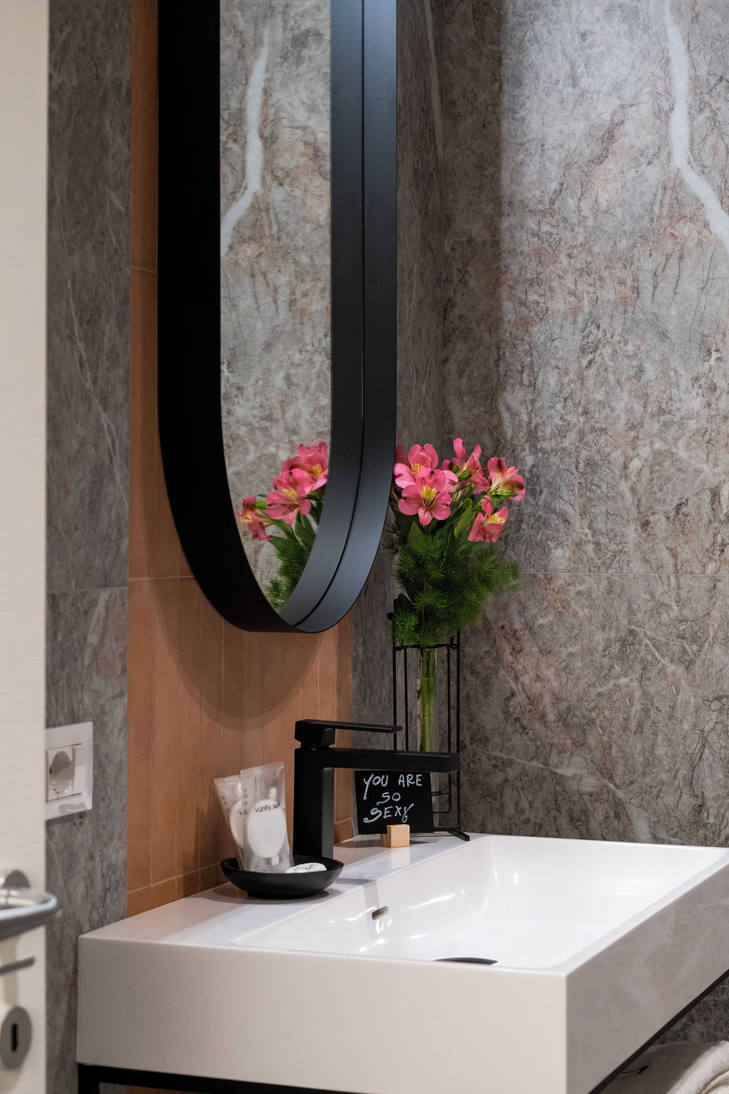 Un bagno con un lavandino bianco, uno specchio nero e fiori rosa.