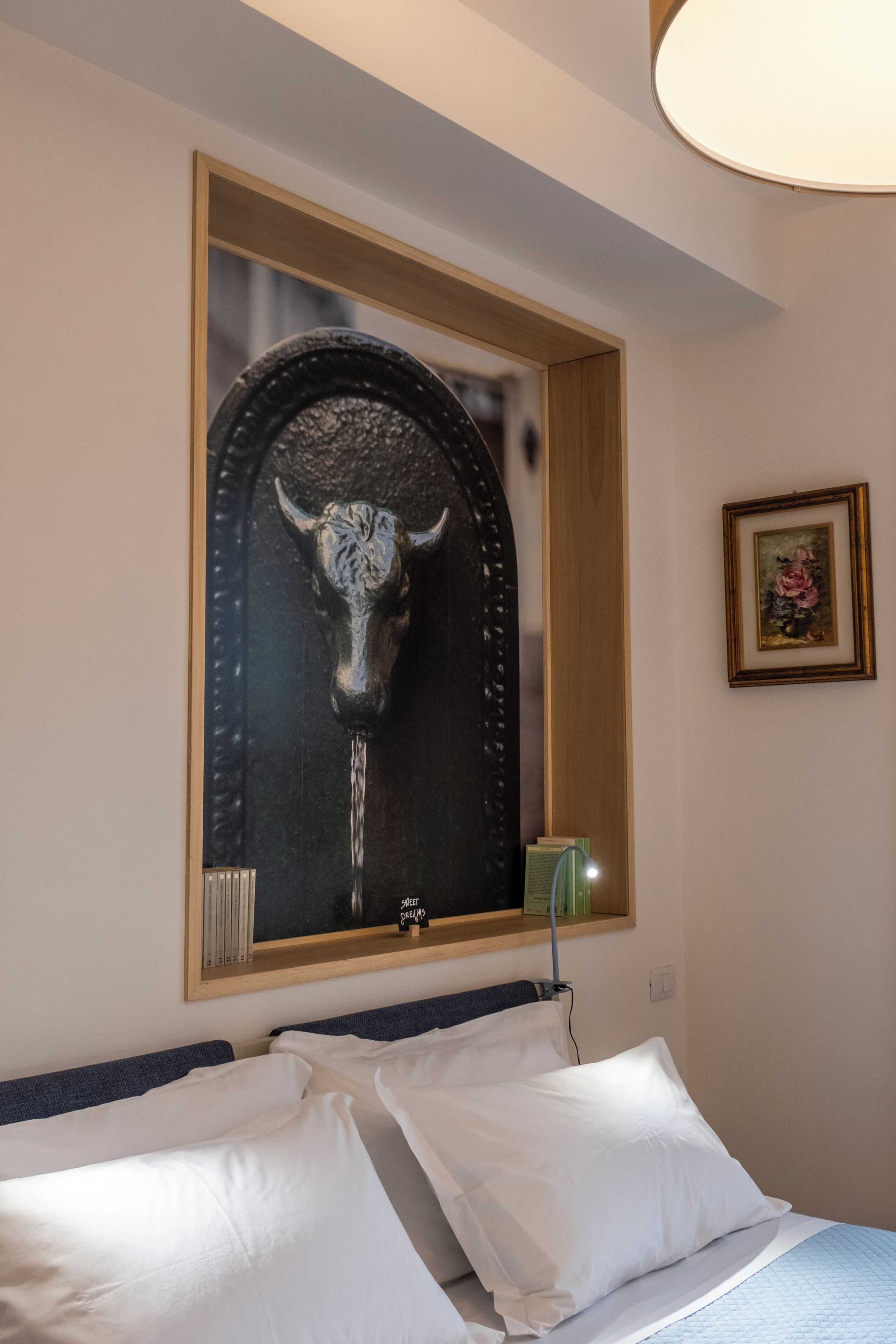 A hotel room with a bed, white pillows, a framed artwork of a bull's head, a small green vase, and a framed picture on the wall.