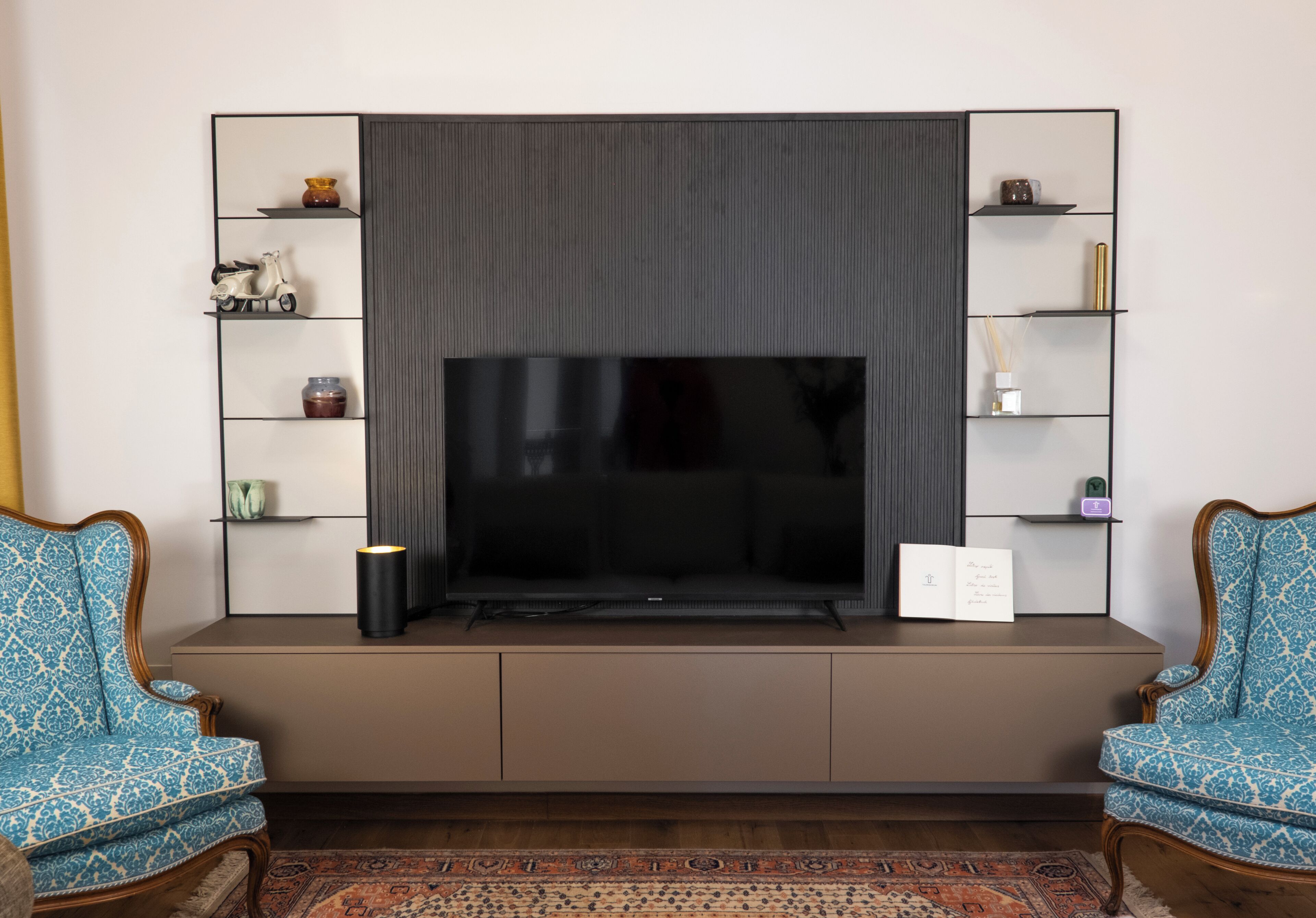 A living room with a large flat-screen TV mounted on a wall, flanked by two blue patterned armchairs and a brown sofa.