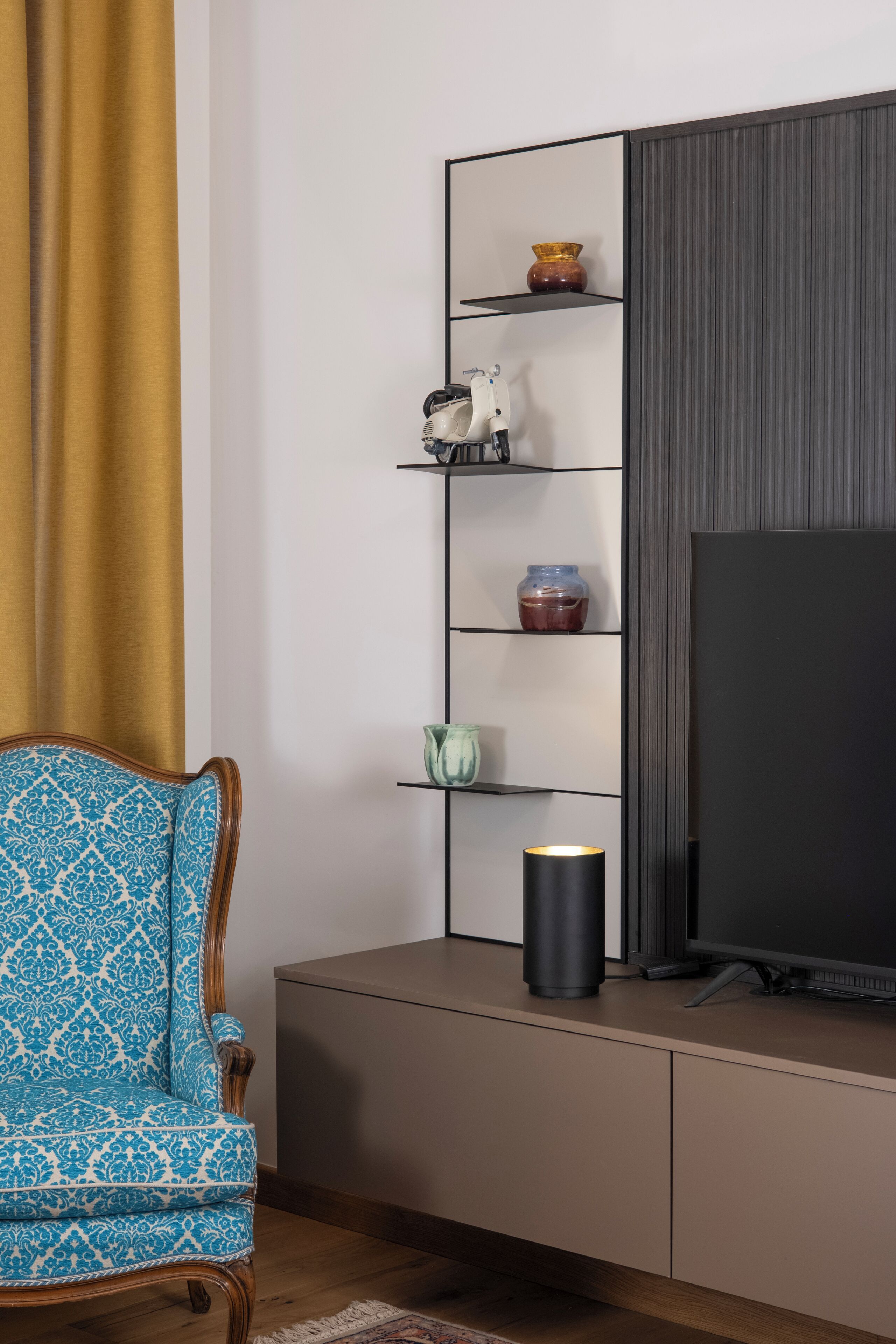 A living room with a TV on a stand, a patterned armchair, a shelf with decorative items, and a curtain.