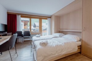 A hotel room with two beds, a desk with a chair, a TV, and a view of the outside. - Charming studio in the center of Saas-Fee (Saas-Fee)