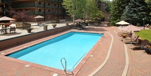 Una piscina rectangular con una escalera metálica, rodeada por un patio de ladrillo. Hay sillas de descanso y sombrillas, y un edificio con balcones al fondo.