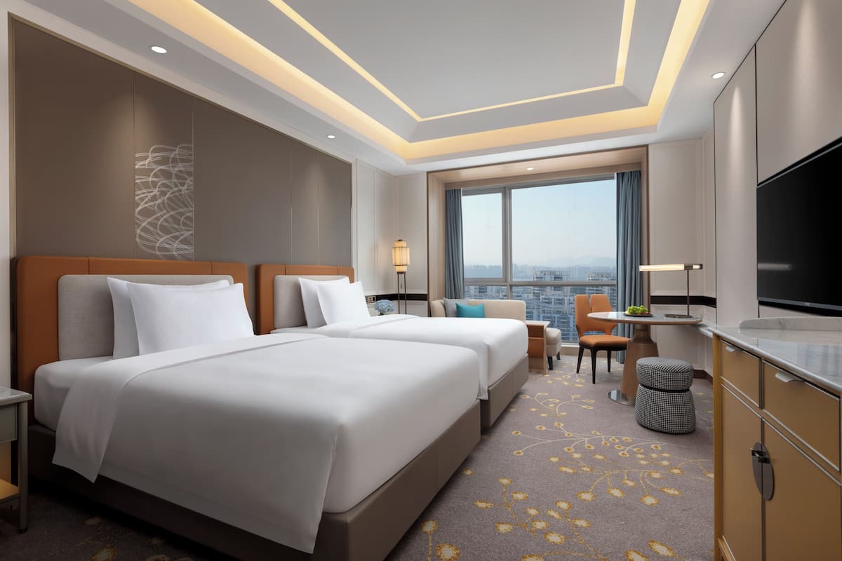 A hotel room with two beds, a TV, a desk with a chair, and a view of the city.