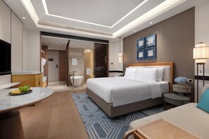 Une chambre d’hôtel moderne dotée d’un grand lit, d’un bureau avec un panier de fruits, d’une salle de bain avec baignoire et d’une télévision.