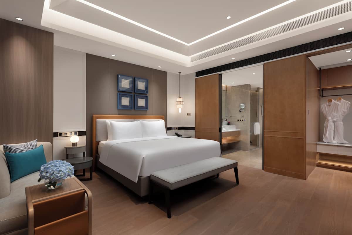 A modern hotel room with a large bed, bedside tables, a bench, a wardrobe, and a bathroom visible through an open door.
