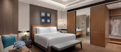 A modern hotel room with a large bed, bedside tables, a bench, a wardrobe, and a bathroom visible through an open door.