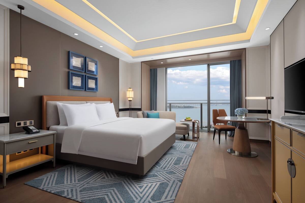 A modern hotel room with a large bed, a desk, a TV, and a view of the city.
