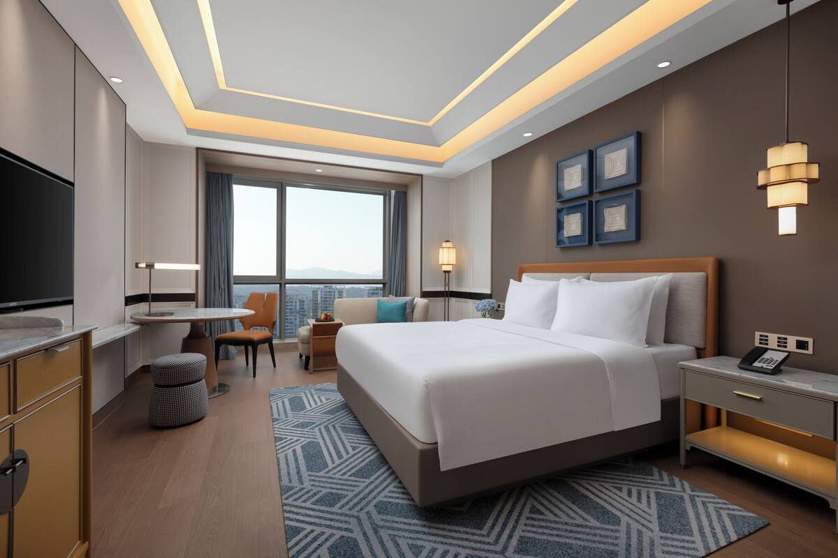 A modern hotel room with a large bed, a desk, a chair, a TV, and a city view.