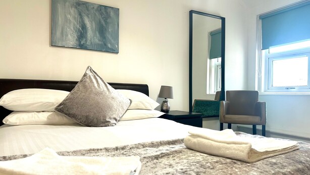 A hotel room with a bed, pillows, a bedside table with a lamp, a chair, a large mirror, and a window.