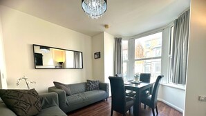 A modern living room with a grey sofa, a dining table with chairs, a chandelier, and a large window with curtains. - Sienna Ground Floor 2Bed with Garden (Blackpool)