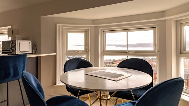 Un coin repas avec une table ronde et quatre chaises, de grandes fenêtres offrant une vue sur la ville, et une kitchenette équipée d’un four à micro-ondes.