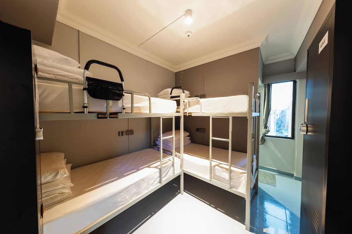 Quarto de hotel compacto com beliches, uma kitchenette pequena e uma janela que oferece luz natural.