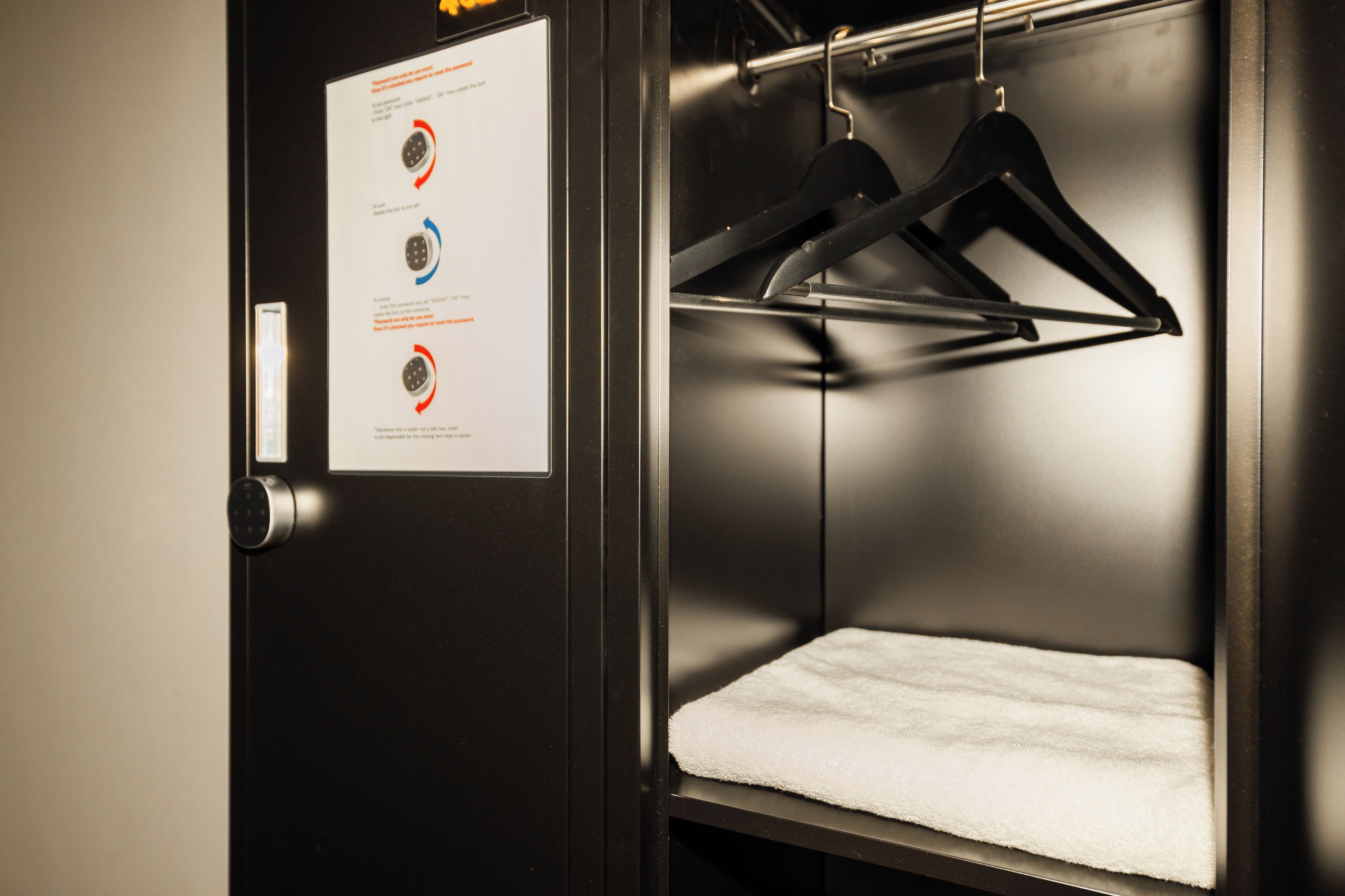 A hotel room with a white towel on the bed, a door with a keypad lock, and a notice board with diagrams.