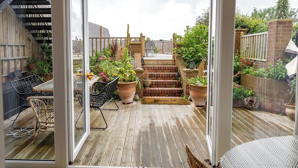 Une terrasse couverte avec une plateforme en bois, des meubles d’extérieur, des plantes en pot et un escalier menant à un niveau supérieur.