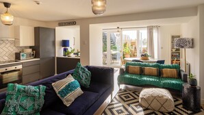 Un salon moderne avec un canapé bleu foncé, des coussins verts et un tapis à motifs. On aperçoit, au fond, une cuisine équipée d’un four à micro-ondes et de placards.