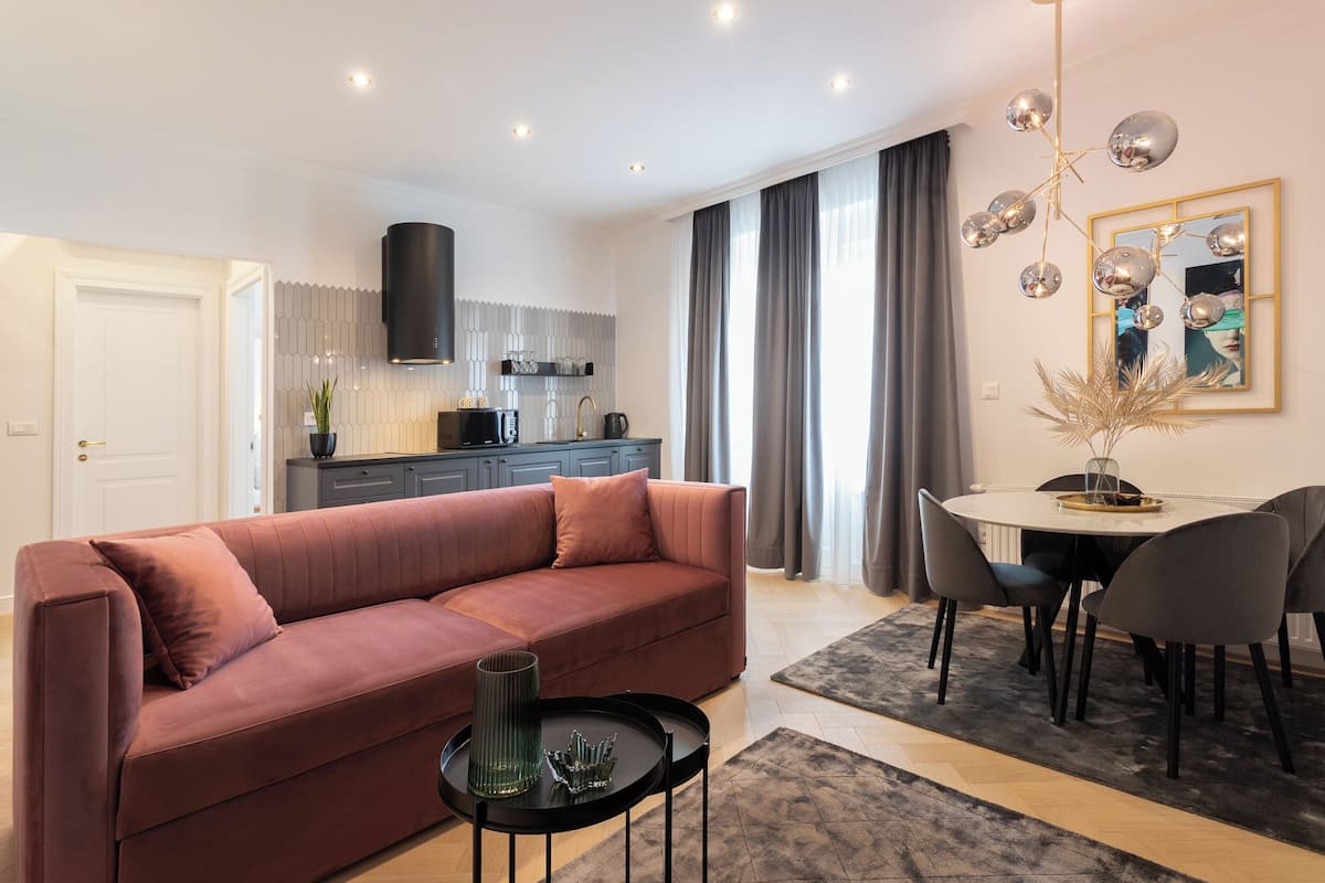 A modern living room with a sofa, a dining table with chairs, a kitchen area, and a large window with curtains.