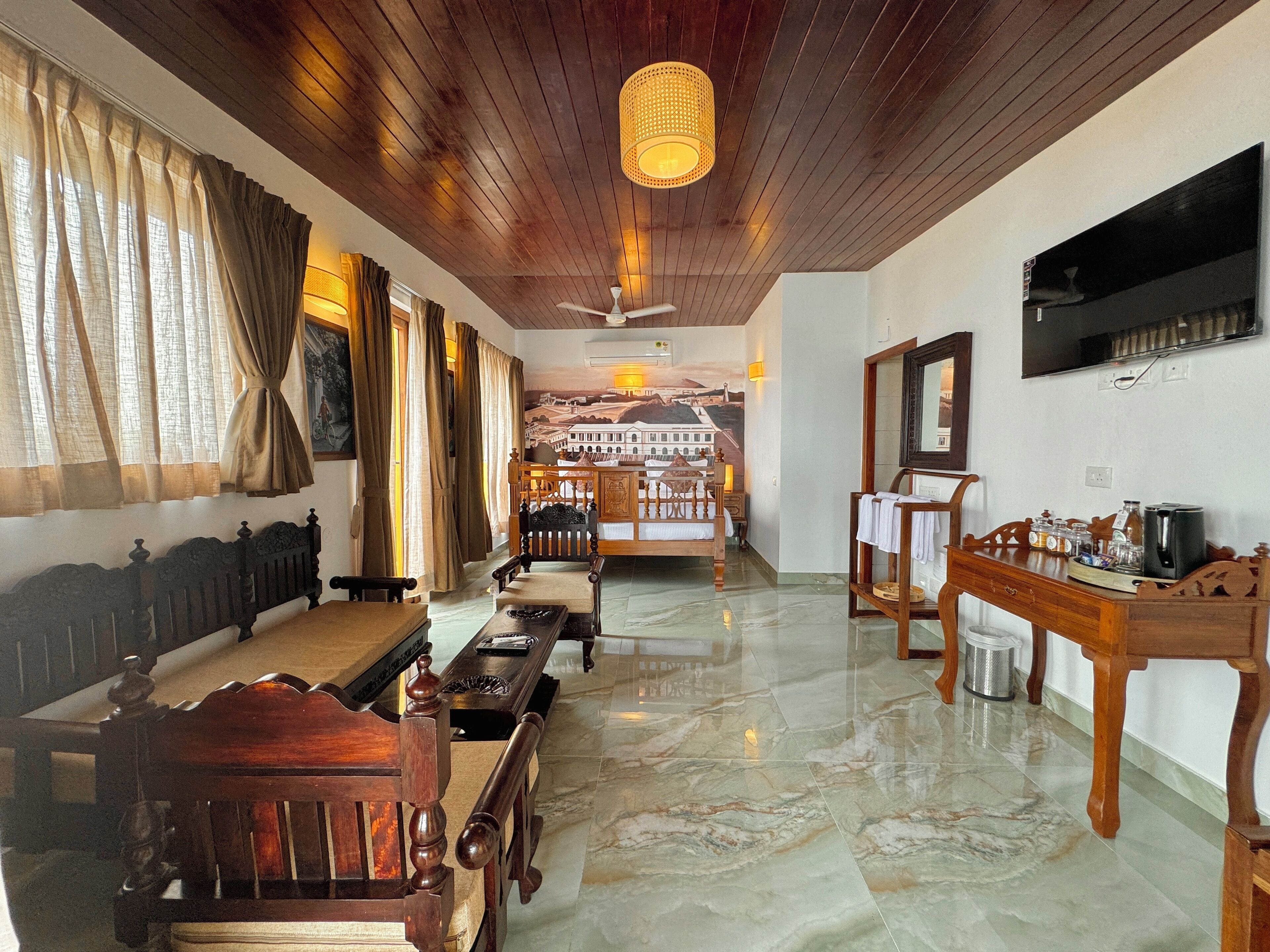 A spacious room with a wooden ceiling, a large window with curtains, a dining area with a table and chairs, a sofa, a television mounted on the wall, and a decorative wall mural in the background.