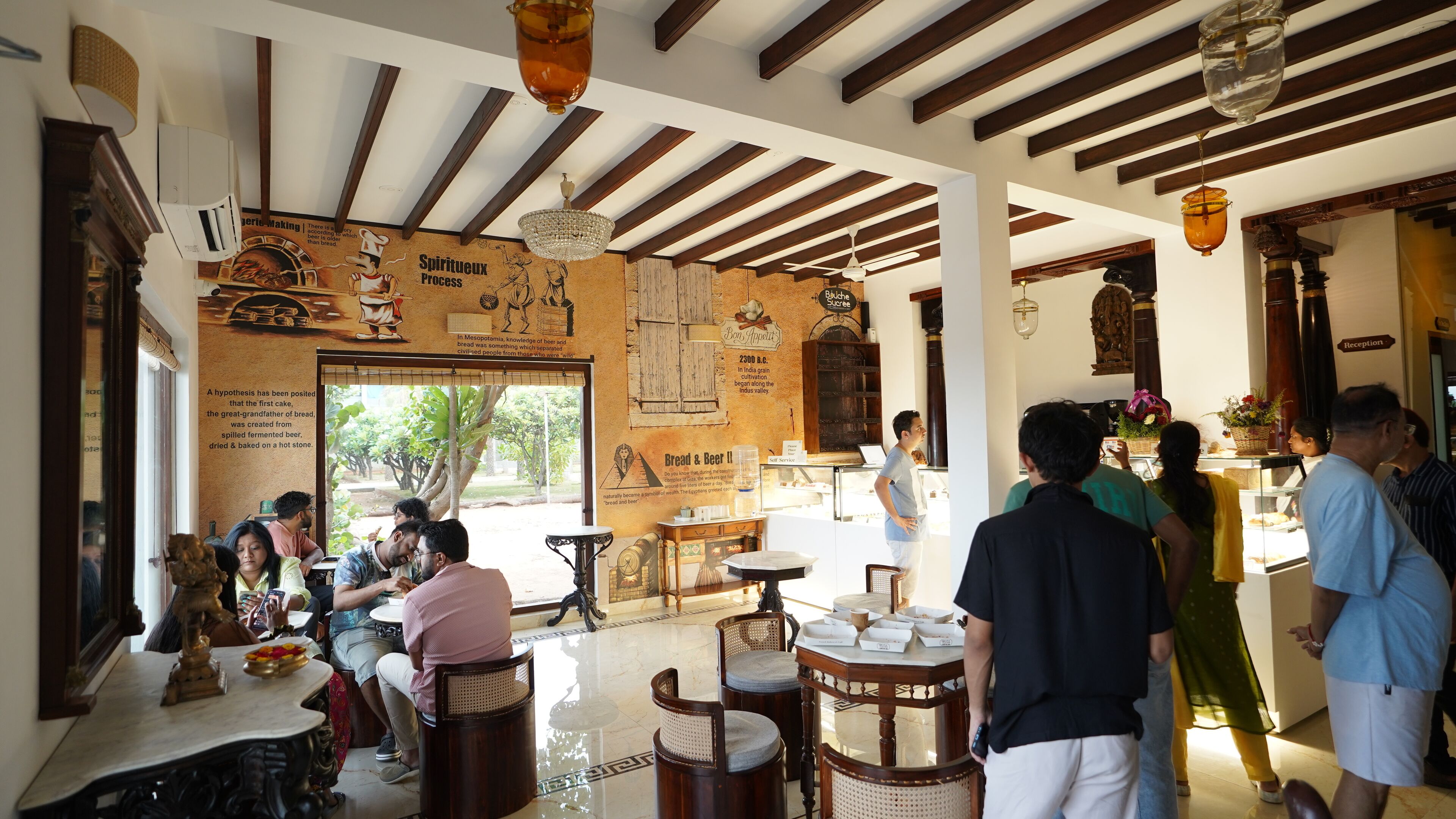 Een restaurant met een hoge plafond, houten balken en een muurschildering van historische figuren en tekst. Er zijn eettafels en stoelen, en op de achtergrond is een receptie te zien.