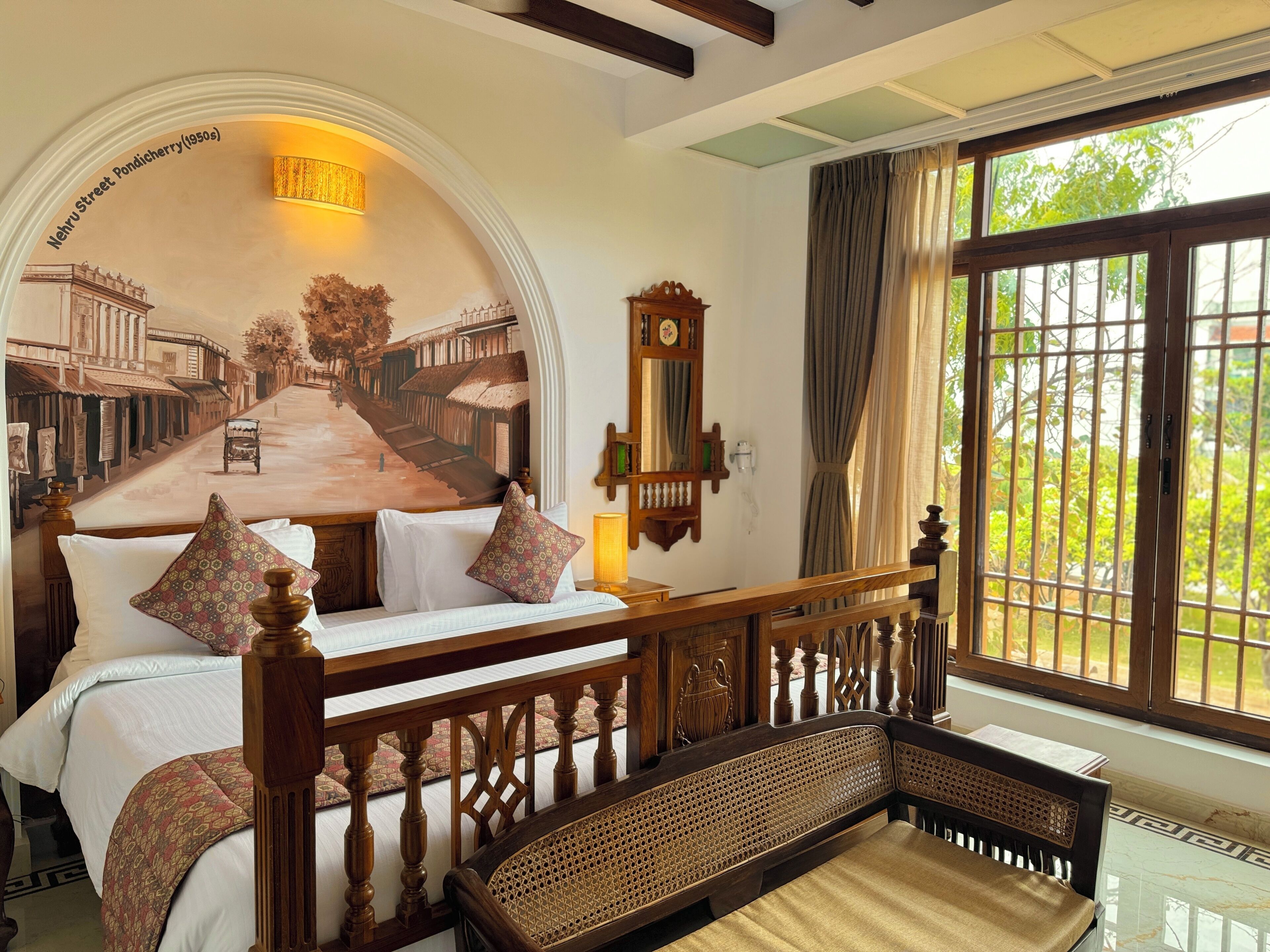 A bedroom with a bed, a bench, a mural, a window, and a mirror.