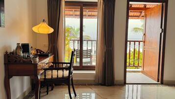 Quarto com escrivaninha de madeira, luminária, cadeira e vista para o oceano através de uma grande janela.