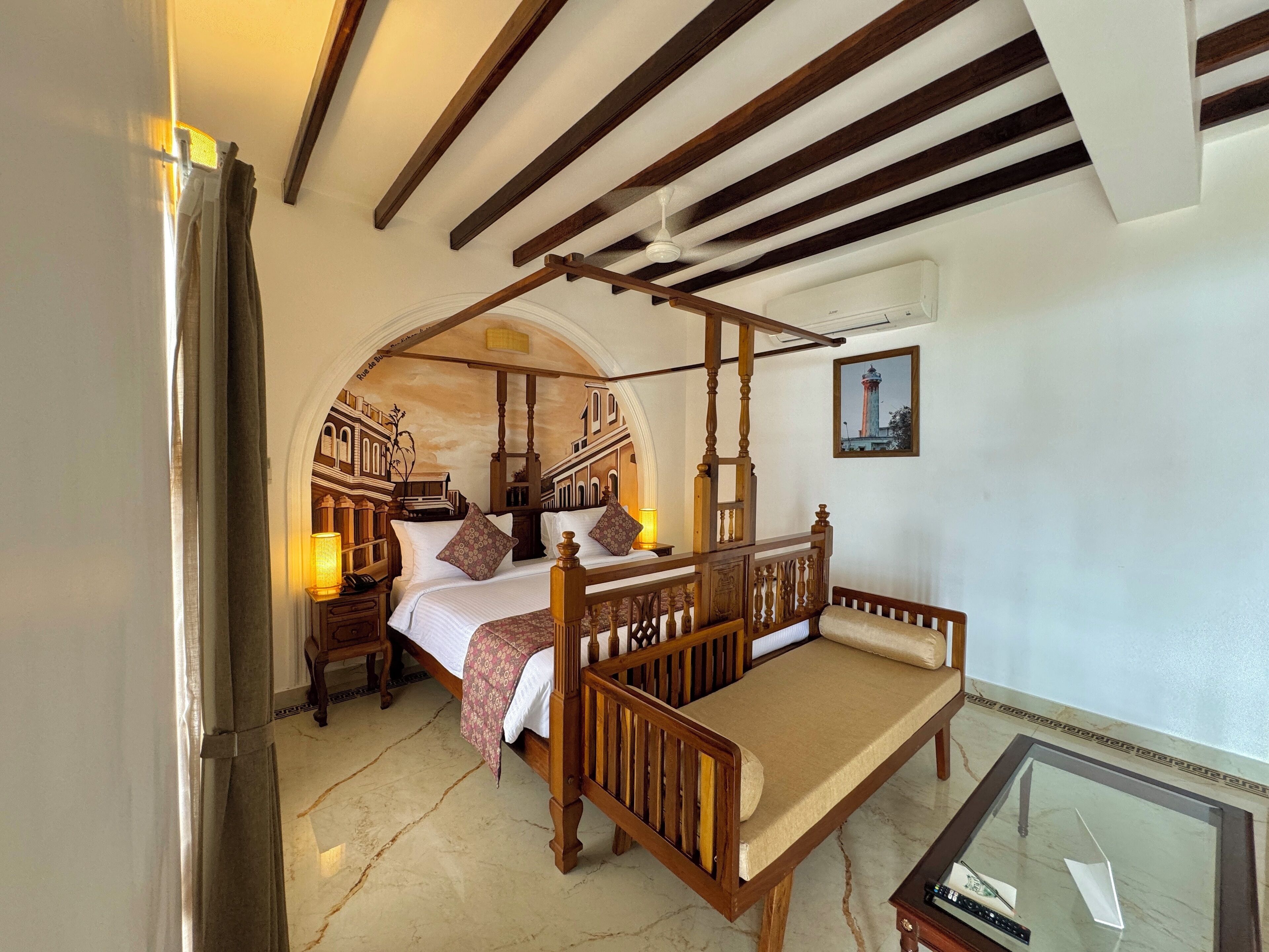 A four-poster bed with a canopy, a wooden bench, a glass table, and a mural of buildings on the wall.
