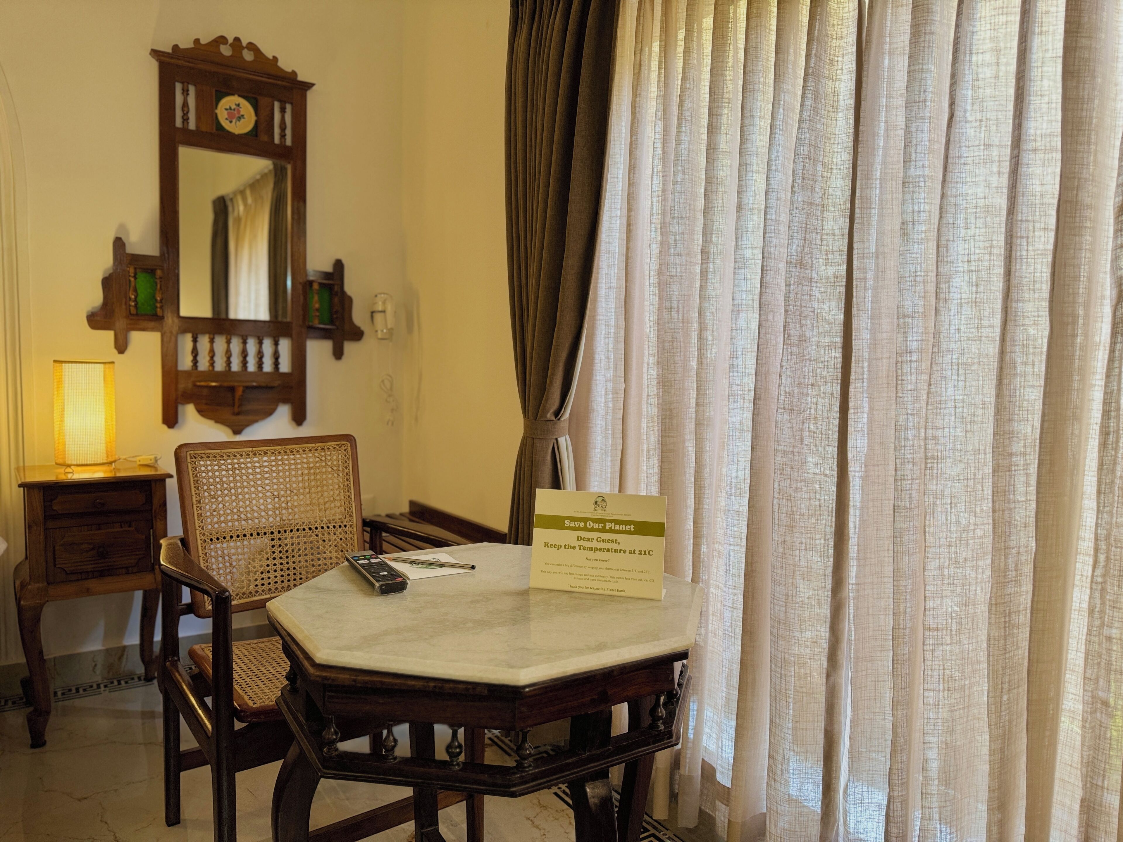A hotel room with a wooden chair, a small table, a lamp, a mirror, and a sign advising to keep the temperature at 21C.