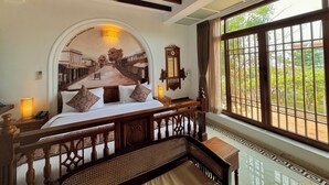 A bedroom with a bed, wooden headboard, a ceiling fan, a large window with a view of greenery, and a mural on the wall. - La Plage SeaView Suites (Puducherry)