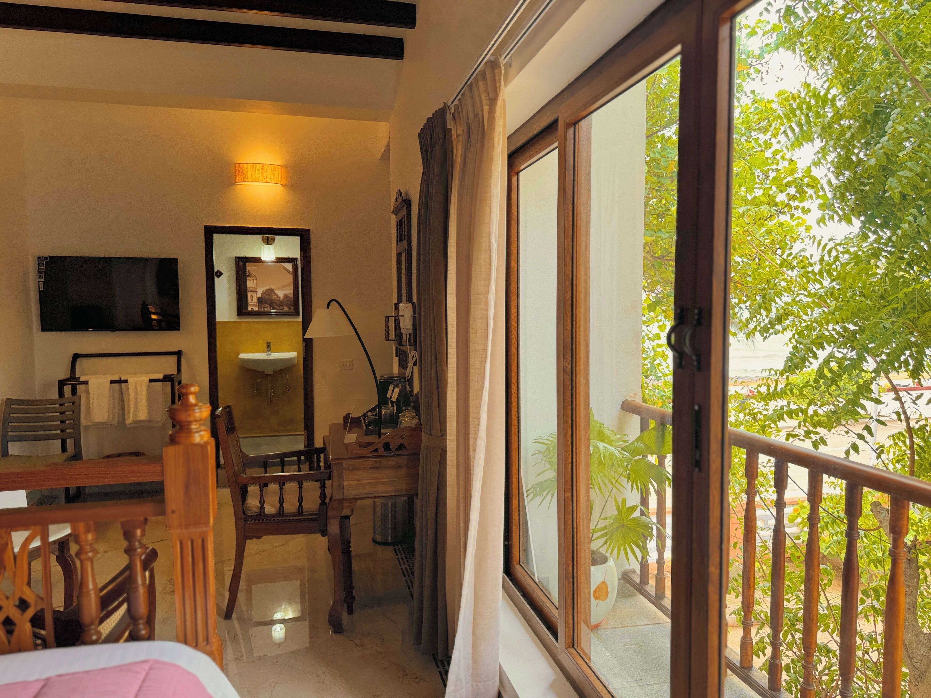 A hotel room with a bed, a desk, a chair, a TV, a mirror, a window with a view of greenery, and a balcony railing.