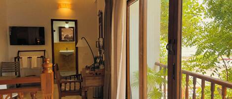 A hotel room with a bed, a desk, a chair, a TV, a mirror, a window with a view of greenery, and a balcony railing.