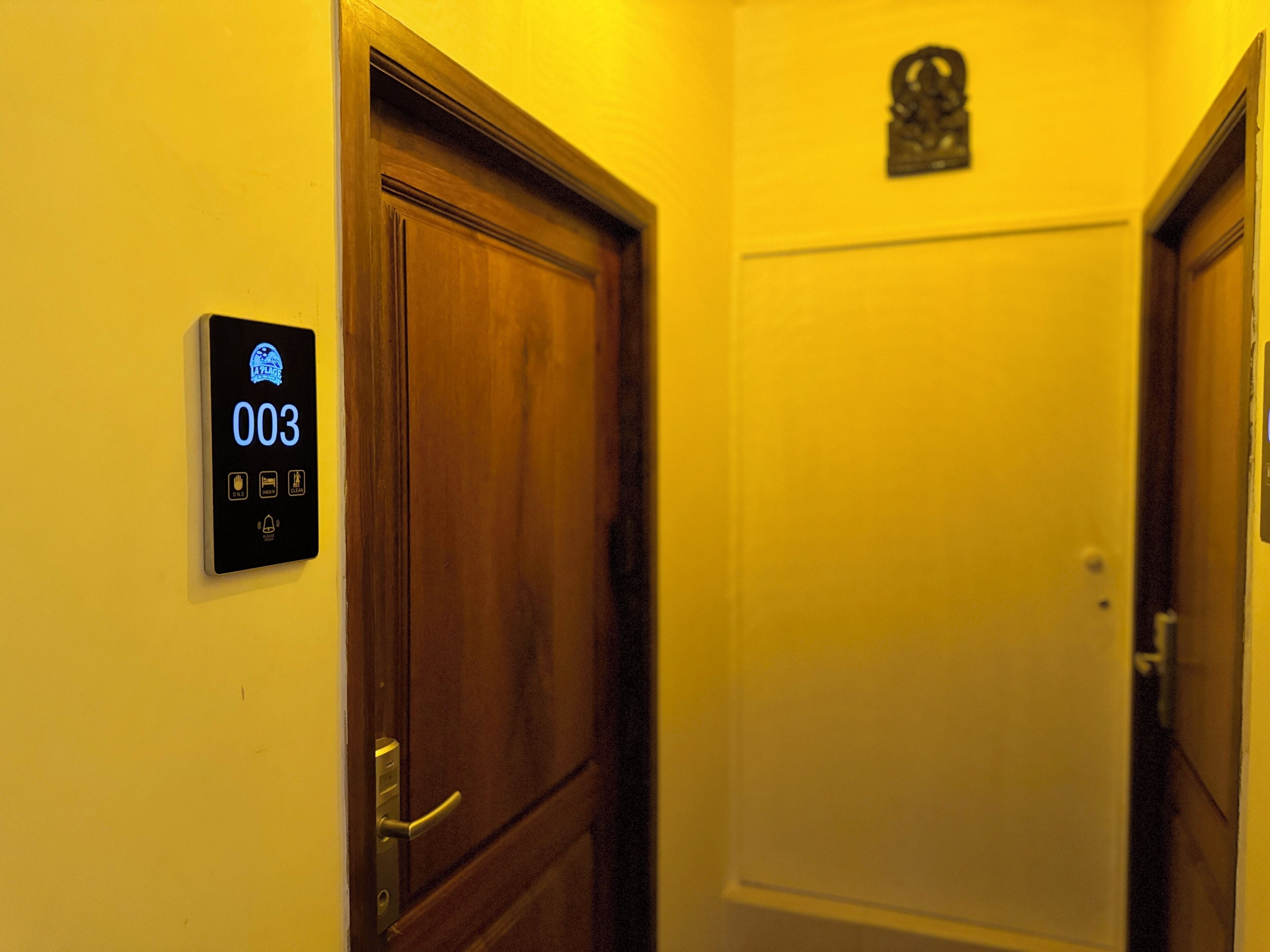 A hotel room with a wooden door, a digital lock panel displaying the number 003, and a small decorative item above the door.